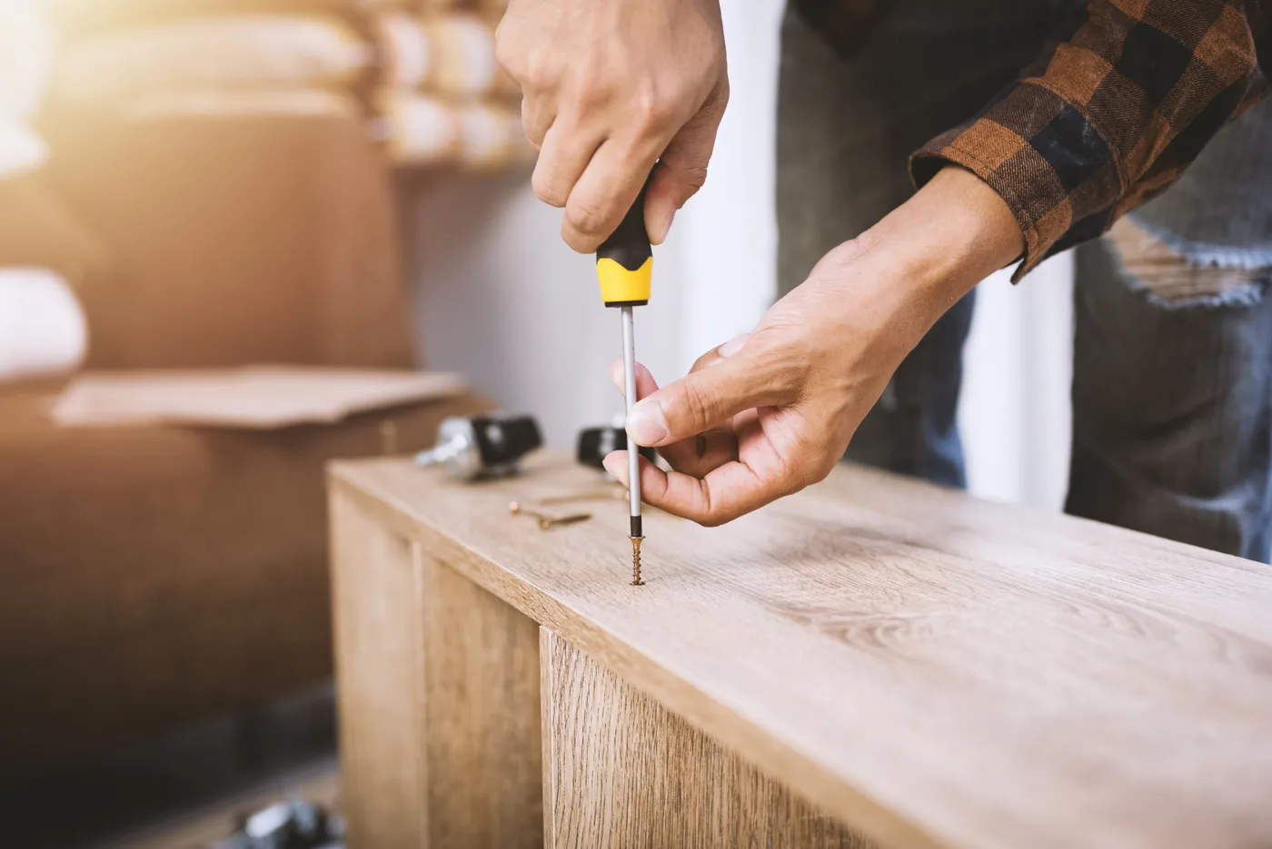 Close-up of a person using a screwdriver to drive a screw into a wooden cabinet, with fittings and packed boxes nearby