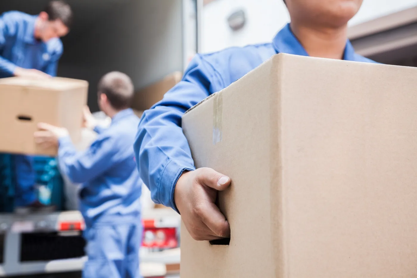 A mover carrying a taped cardboard box past an open truck while two colleagues load behind