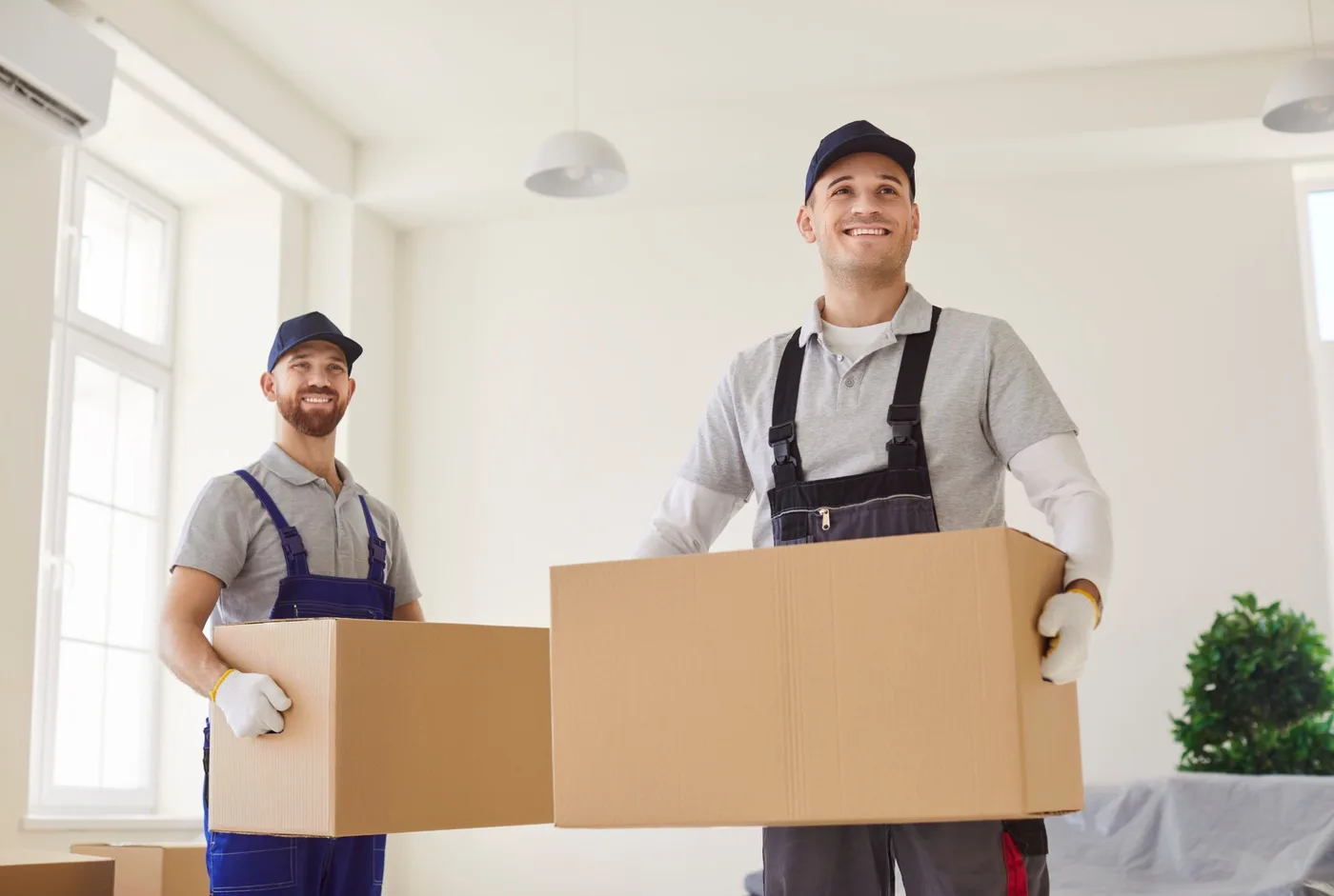 Two movers carrying cardboard boxes through a bright, empty room with bare walls
