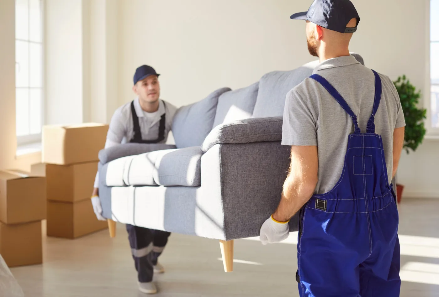 Two movers carrying a grey sofa together through a bright, empty room