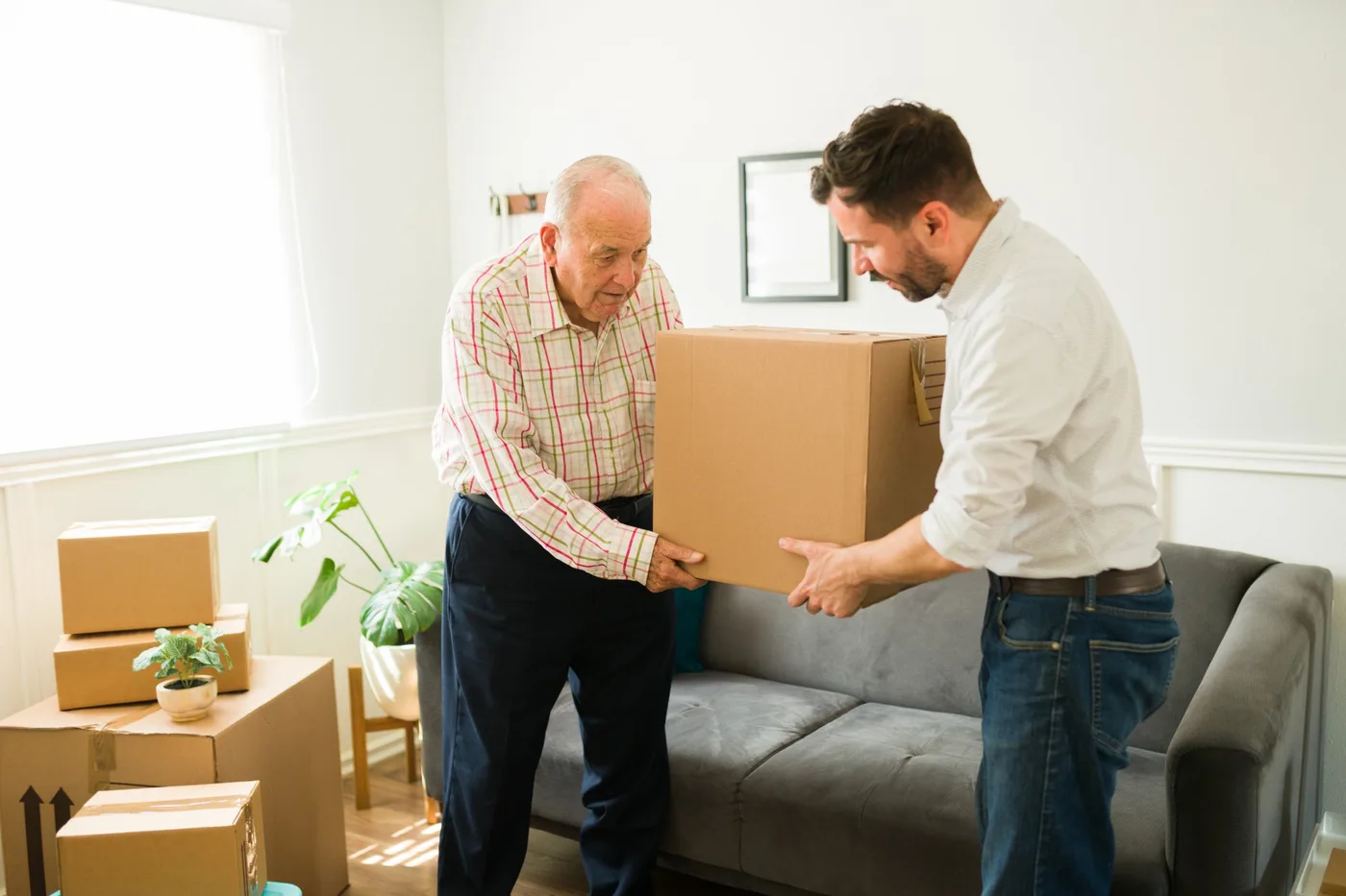An older man handing a cardboard box to a younger man in a living room, with packed boxes and a sofa around them