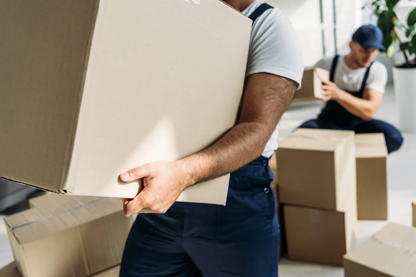 A mover carrying a large cardboard box while a colleague packs another box behind him