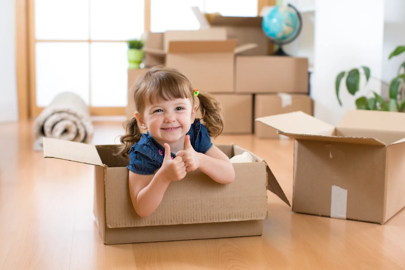 A smiling toddler sitting inside an open cardboard box giving two thumbs up, in a room with packed boxes and a rolled rug