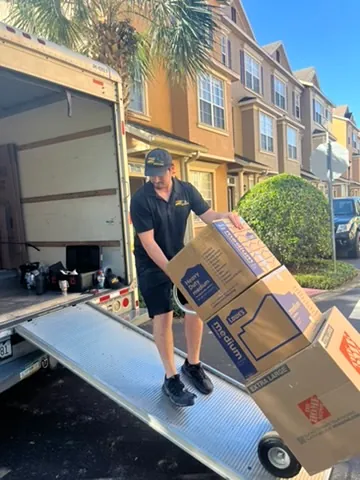 Man wearing black moving boxes on a dolly down a ramp from a moving truck in a residential area.