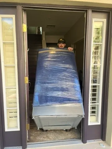 Man wearing a cap moving a large wrapped piece of furniture or appliance through a doorway with stairs visible inside.