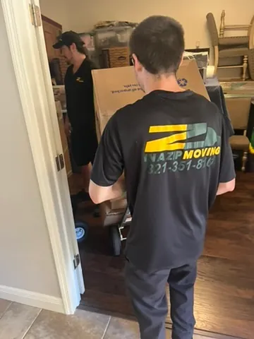 Two movers in black uniforms moving a large cardboard box inside a home with wooden floors and stacked chairs.