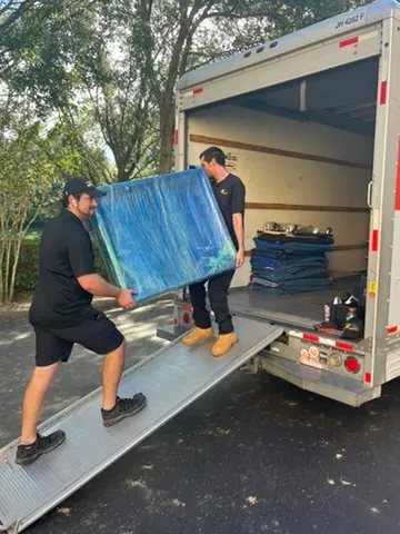 Two movers carrying a large wrapped item on a ramp into a moving truck during daytime.